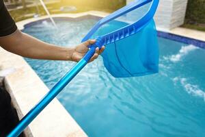 persona con desnatadora limpieza piscina, manos participación un desnatadora con azul piscina en el antecedentes. hombre limpieza el piscina con el desnatadora, un hombre limpieza piscina con hoja desnatadora foto