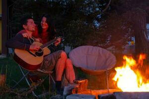 un hombre obras de teatro el guitarra, un mujer escucha y canta a lo largo de. un Pareja en amor es sentado por el al aire libre hoguera en el patio de el casa en cámping sillas, un romántico noche foto