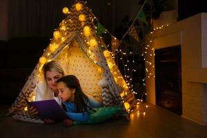 madre y hija son sentado en un tipi carpa, leyendo cuentos con el Linterna. contento familia. foto