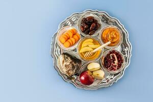 un plato con un nuevo años tratar para el nuevo años fiesta rosh hashaná. granada, Miel, fechas, manzana y zanahorias. azul antecedentes. Copiar espacio. foto