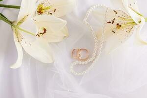 Beautiful wedding background with a pair of wedding gold rings, pearl beads and white live lilies. fabric satin white background. photo