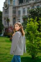 Girl on city walk. Young woman standing with her back in half turn and looking directly at camera. Cool weather in summer, girl draped jacket over her shoulders. Old building and trees on long plan. photo