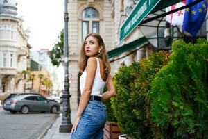 Young woman standing in half-turn with her back on sidewalk. Historic building. Beautiful girl wearing casual clothes and looking into distance. Batumi Historic Site in Georgia. Blurred background photo