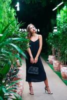 Glamorous model wearing black long strappy dress with her bag in her hand. Girl standing among tropical green plants inside arrangement. Thinking sad beautiful woman. photo