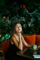 hermosa joven mujer emplazamiento en suave sofá a mesa en cafetería. pensamiento niña mirando dentro distancia. verde plantas en antecedentes. caliente bebida taza en mesa. joven mujer vistiendo negro vestir en Delgado correas foto