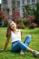 hermosa joven mujer vistiendo tanque parte superior y pantalones sentado en césped césped. verde césped en frente de antiguo edificio. soleado día en parque, verde césped y arboles en antecedentes. calma niña disfrutar relajante en ciudad. foto