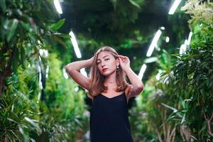 Young woman wearing black dress on thin straps stands among green plants inside orangery. Fashionable girl posing in greenhouse or botanical garden. Young female model in evening outfit photo