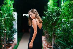 Beautiful woman standing with her back among tropical potted plants. Young woman wearing black dress dress with open back inside flower greenhouse. Girl with long hair. Blurred background photo