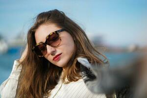 Close up image of brunette woman wearing stylish sunglasses posing outdoors. Long-haired Girl with bright makeup enjoying vacation. Beautiful girl standing against backdrop of sea photo