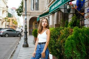 Empowered young woman standing on sidewalk. Long-haired brunette wearing blue jeans and white tank top. City Walk in Historic District. photo