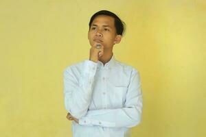 a man in white stood with his hand on his chin showing a thoughtful expression.  man with confused face isolated in yellow backgroumd photo