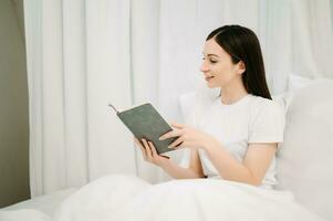 retrato de bueno sano joven mujer leyendo libro y descansando en cama a dormitorio. estilo de vida concepto foto