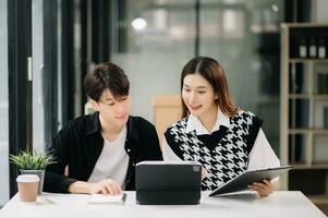 Asian business colleague business presents and explains and using laptop and tablet. Teamwork, financial marketing team, while sitting in modern office room. photo