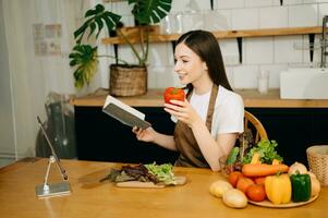 Young beautiful woman in the kitchen in an apron, fresh vegetables on the table, writes down her favorite recipes, comes up with ideas for dishes photo