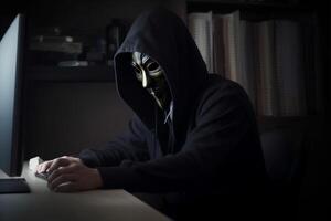 Anonymous hacker with hood and mask sitting next to computer photo