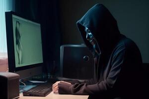 Anonymous hacker with hood and mask sitting next to computer photo