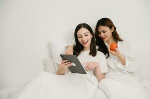 Smiling young sisters lovely couple sitting on white bed together with tablet, smartphone and laptop in bedroom. Homosexual women or Lesbian concept. photo