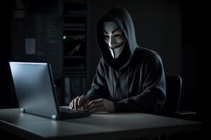 Anonymous hacker with hood and mask sitting next to computer photo