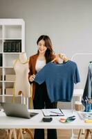 asiático joven mujer en escritorio en oficina de Moda diseñador y sostiene tableta, ordenador portátil y teléfono inteligente en blanco mesa foto