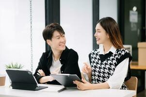 Asian business colleague business presents and explains and using laptop and tablet. Teamwork, financial marketing team, while sitting in modern office room. photo