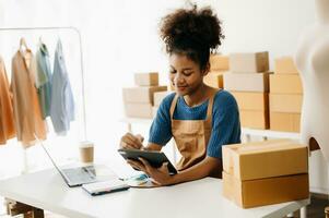 puesta en marcha pequeño negocio pequeño, empresario propietario africano mujer utilizando teléfono inteligente o tableta tomando recibir y comprobación en línea compra compras orden a preparando paquete producto caja. en oficina foto