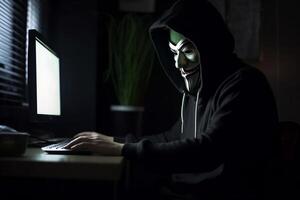 Anonymous hacker with hood and mask sitting next to computer photo