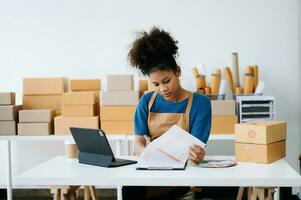 Startup small business SME, Entrepreneur owner African woman using smartphone or tablet taking receive and checking online purchase shopping order to preparing pack product box. in office photo