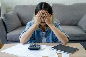 Asian woman sitting covering face with hands with many bills on table at home. she feels stressed. Debt or bankruptcy concept. photo