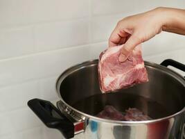 lady putting raw pork chop in metal pot for preparing soup in white clean home kitchen, home cooking concept photo