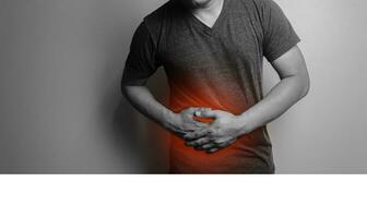 Close up black and white image a man with a gray T-shirt with a hand on his stomach because of indigestion, feeling sick appendicitis Pain concept. Stands on gray background. photo