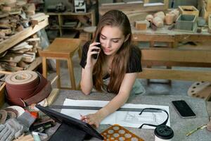 Cheerful young carpenter discussing order details with her customer on smartphone and taking necessary notes, interior of spacious workshop on background photo