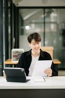 Young attractive Asian man smiling thinking planning writing in notebook, tablet and laptop working from home, looking at camera at office photo