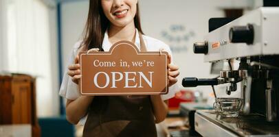 Young female entrepreneur hanging a welcome sign in front of a coffee shop. Beautiful waitress or hostess holding a tablet preparing photo