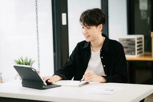 Young attractive Asian man smiling thinking planning writing in notebook, tablet and laptop working from home, looking at camera at office photo