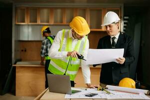 Contractor and architect looking at plan, tablet, laptop and talking about working on big project. Building in construction interior. photo