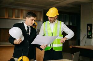 Contractor and architect looking at plan, tablet, laptop and talking about working on big project. Building in construction interior. photo