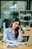 mujer asiática de negocios hablando por teléfono y usando una laptop con una sonrisa mientras está sentada en la oficina foto