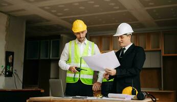Contractor and architect looking at plan, tablet, laptop and talking about working on big project. Building in construction interior. photo