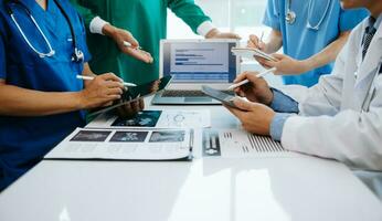 Medical team having a meeting with doctors in white lab coats and surgical scrubs seated at a table discussing a patients working online using computers in the medical industry photo