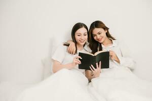 Smiling young sisters lovely couple sitting on white bed together with tablet, smartphone and laptop in bedroom. Homosexual women or Lesbian concept. photo