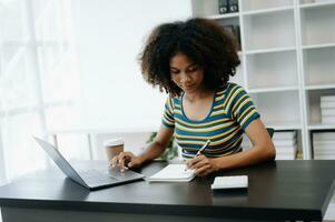 Beautiful African woman using laptop and tablet while sitting at her working place. Concentrated at work in home office photo