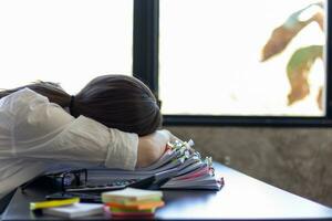 secretary was laying her head down on stacked documents as she was tired searching through huge stacked documents before meeting. difficulty in finding hire purchase contract from stacked documents photo