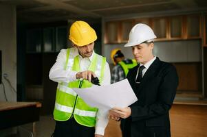 Contractor and architect looking at plan, tablet, laptop and talking about working on big project. Building in construction interior. photo