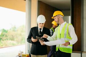 Contractor and architect looking at plan, tablet, laptop and talking about working on big project. Building in construction process photo