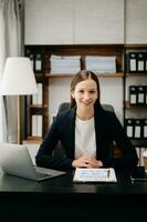 Confident business expert attractive smiling young woman typing laptop ang holding digital tablet on desk in office. photo