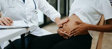 Pregnant african woman has appointment with doctor at clinic. Male gynaecologist OB GYN medic specialist with stethoscope listens to baby's heartbeat in mother's belly. Pregnancy, health care photo