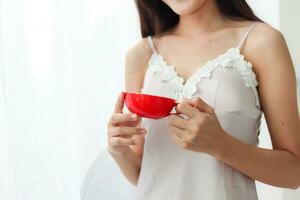 A young woman wearing a sexy white see through nightgown stands by the window with a red cup of coffee in the morning to drink coffee and see the beautiful scenery of the sunrise in the morning. photo