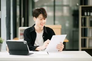 joven atractivo asiático hombre sonriente pensando planificación escritura en computadora portátil, tableta y ordenador portátil trabajando desde hogar, mirando a cámara a oficina foto