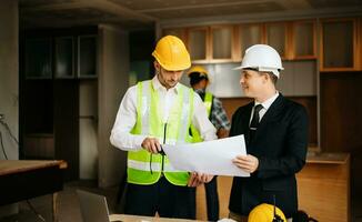 Contractor and architect looking at plan, tablet, laptop and talking about working on big project. Building in construction interior. photo