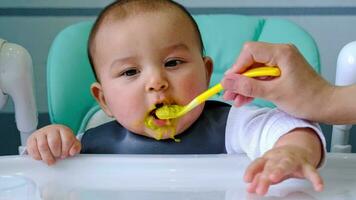 A dirty, joyful baby at the table on a feeding chair got his mouth dirty in vegetable puree and knocks his hand. Introduction of complementary foods, the child likes to eat. Portrait close-up video